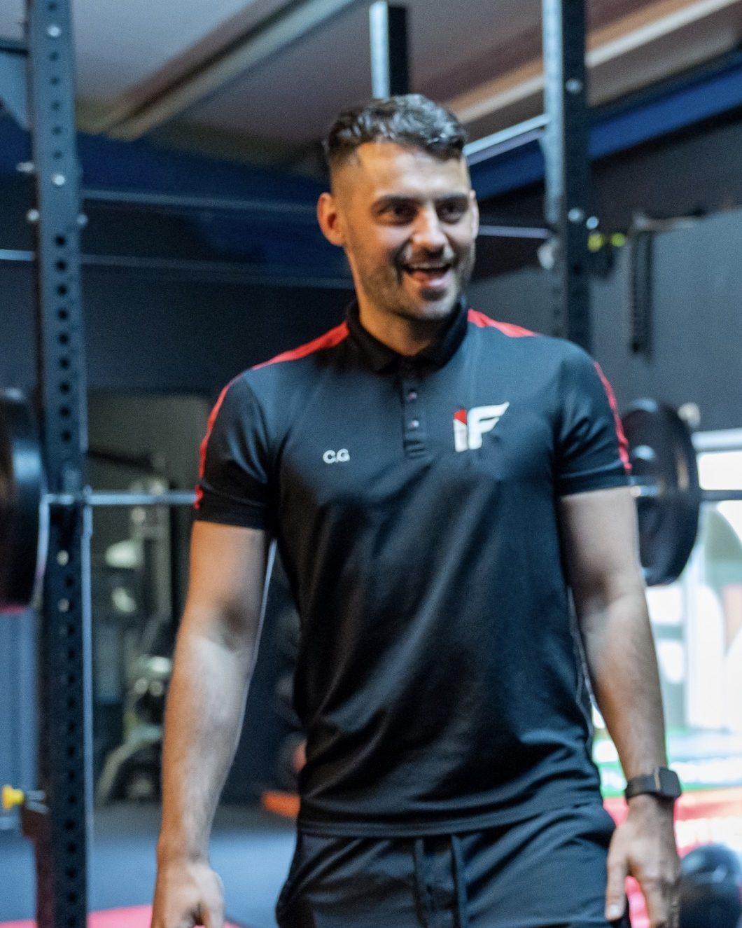 Gibby, head coach at Intensity Fitness, smiling in the gym in a black and red IF polo shirt